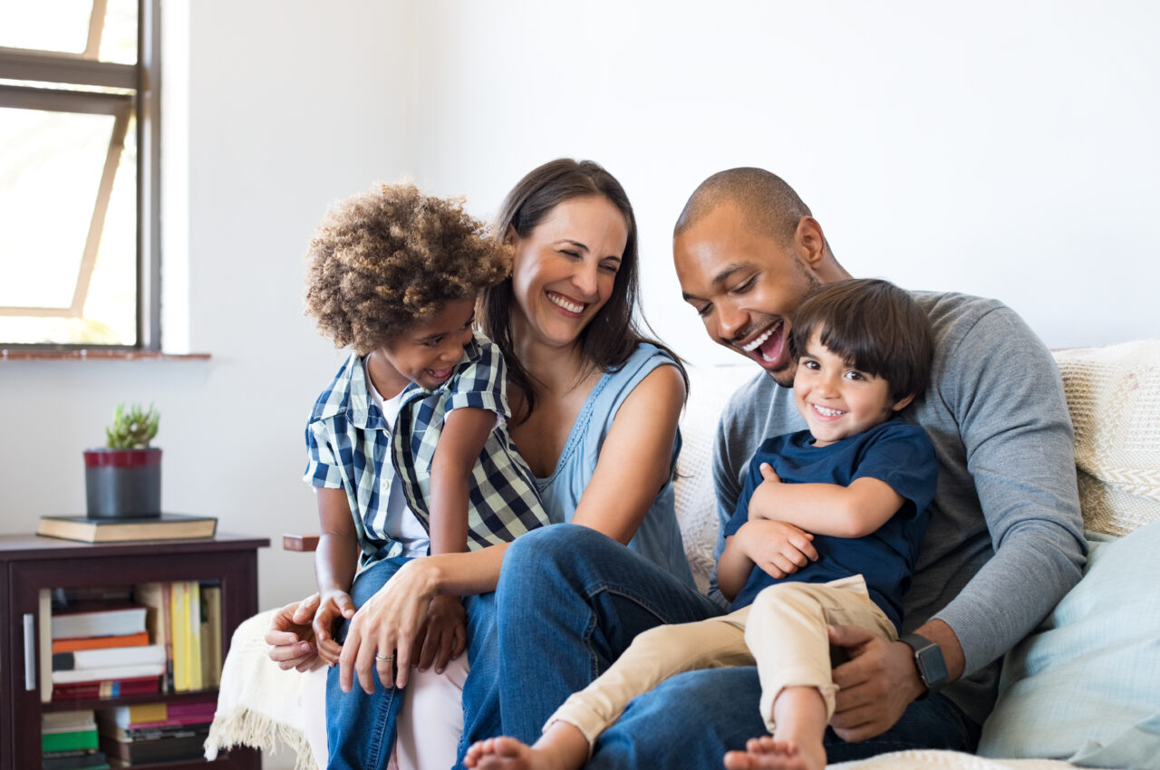 Happy family sitting on sofa laughing together. Cheerful parents playing with their sons at home. Black father tickles his little boy while the mother and the brother smile.