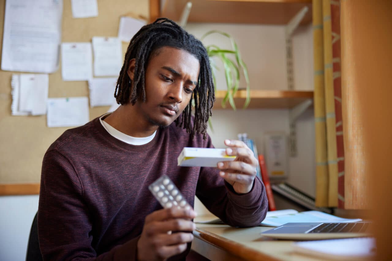 University Or College Student With Poor Mental Health Looking At Anti Depressant Medication Packaging At Desk In Room