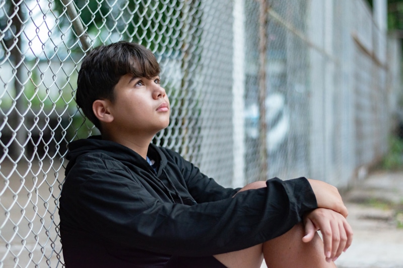Asian teenager sitting alongside a fence looking to the sky in search of something.